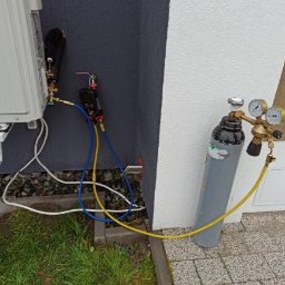 AGO Tomasz Stawowy - Podłączenie butli z gazem do zewnętrznej jednostki klimatyzacji podczas instalacji, widoczne przewody pomiarowe i manometry.