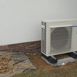 AGO Tomasz Stawowy - Zewnętrzna jednostka klimatyzacji Panasonic na betonowej podstawie, zainstalowana przy ścianie domu w Rzykach, obok widoczna sterta ziemi i materiały budowlane.