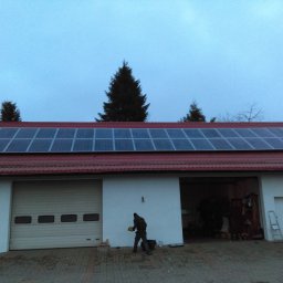 AGO Tomasz Stawowy - Montaż paneli fotowoltaicznych na dachu budynku gospodarczego z czerwoną dachówką, widoczny pracownik w trakcie pracy, pochmurne niebo.