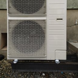 AGO Tomasz Stawowy - Zewnętrzna jednostka klimatyzacji zamontowana na czarnej, stalowej konstrukcji wsporczej, z widocznymi śladami wilgoci, na tle betonowej ściany z pęknięciem i żwirowego podłoża.