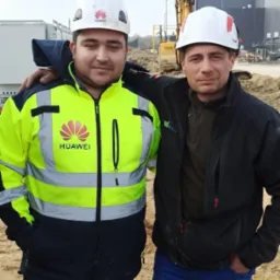 Dwóch inżynierów w kaskach i odzieży roboczej, z logo Huawei na kasku i kurtce, pozuje na placu budowy z koparką w tle, prawdopodobnie po zakończonej realizacji inwestycji.
