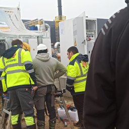 AGO Tomasz Stawowy - Trzech monterów w odblaskowych kamizelkach Huawei instaluje moduł magazynu energii na betonowych fundamentach w terenie budowy, widoczny częściowo czarny budynek w tle.