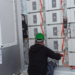 AGO Tomasz Stawowy - Pracownik w zielonym kasku instaluje moduły w szafie magazynu energii, widoczne pomarańczowe pasy mocujące i miernik cęgowy na jednym z modułów.