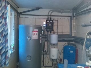 Szara pionowa obudowa zasobnika ciepłej wody użytkowej w kotłowni, z widocznymi rurami, zaworami i białymi zbiornikami wyrównawczymi na tle wykafelkowanej ściany. Okno z zasłoną po lewej stronie.