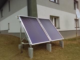 Dwa panele solarne zamontowane na betonowych blokach przed budynkiem z szarą elewacją i anteną satelitarną.