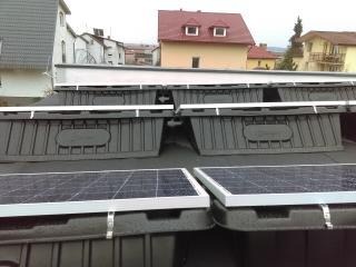 Baterie słoneczne zamontowane na płaskim dachu, umieszczone w czarnych, plastikowych obudowach z białymi elementami mocującymi, widok z bliska na tle zabudowy jednorodzinnej.