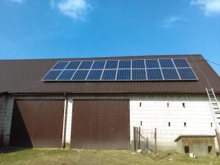 Panele fotowoltaiczne zamontowane na dachu budynku gospodarczego z dwoma bramami garażowymi, drabina oparta o ścianę, błękitne niebo.