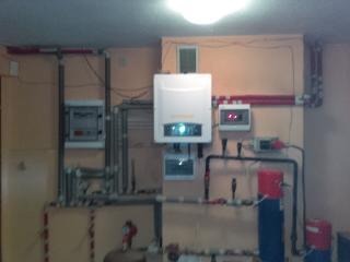 Kotłownia z białym piecem wiszącym, rozbudowaną instalacją hydrauliczną z rurami miedzianymi i plastikowymi, zbiornikami wyrównawczymi oraz skrzynką sterującą na ścianie w pomieszczeniu o beżowych...