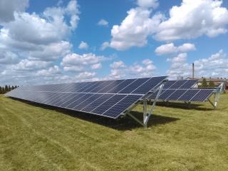 Rozległa instalacja fotowoltaiczna na polanie, panele ustawione rzędami na metalowych konstrukcjach, błękitne niebo z obłokami w tle.