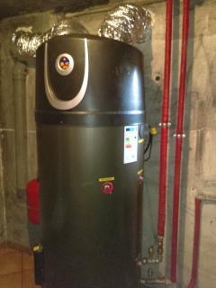 Czarny zbiornik ciepłej wody użytkowej z podłączonymi rurami miedzianymi i elastycznymi przewodami wentylacyjnymi, zamontowany w pomieszczeniu z surowymi, betonowymi ścianami.