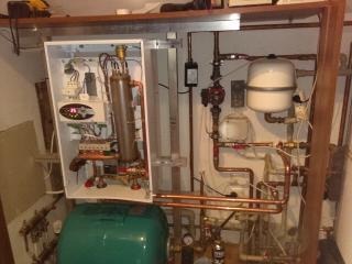 Otwarty panel sterowania kotła grzewczego z widocznymi elementami instalacji, rurami miedzianymi, zbiornikiem wyrównawczym i osprzętem w kotłowni.