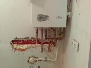 Biały piec gazowy zamontowany na ścianie z widocznymi rurami miedzianymi, częściowo ukrytymi w ścianie z czerwoną izolacją, oraz wystającą białą rurą z pianką montażową u dołu. Widoczny stary...