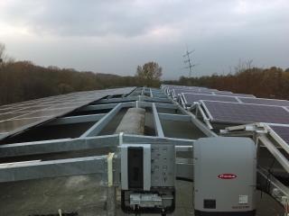 Instalacja paneli fotowoltaicznych na płaskim dachu z widocznym inwerterem marki Fronius. Metalowa konstrukcja wsporcza, pochmurne niebo w tle.