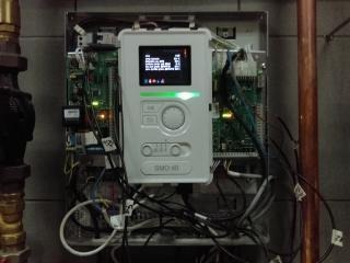 Panel sterowania systemu grzewczego, model SMO 40, zamontowany na ścianie, widoczne okablowanie i elementy instalacji, rury miedziane w tle.