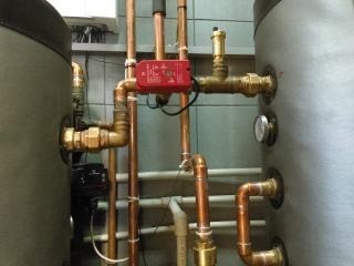 Miedziane rury instalacji grzewczej z czerwonym zaworem i manometrem, widoczne zbiorniki akumulacyjne ciepła w szarej izolacji.