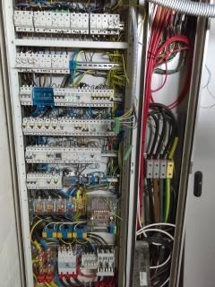 Otwarta szafa sterownicza z widocznymi licznymi bezpiecznikami, przekaźnikami, przewodami w różnych kolorach i listwami zaciskowymi. Część przewodów ułożona w peszlu ochronnym.