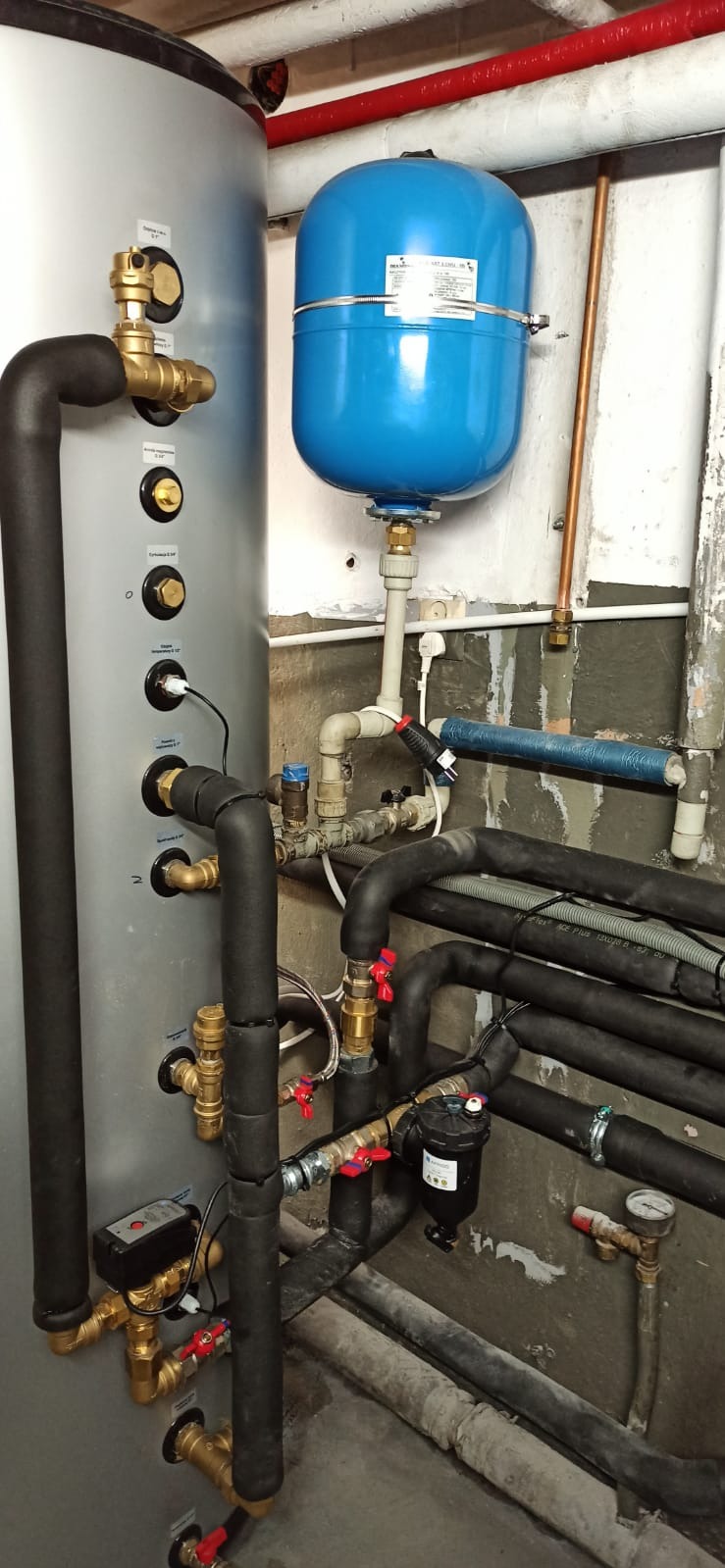 Szary zbiornik buforowy z licznymi rurami, zaworami i czarną izolacją, niebieski zbiornik wyrównawczy ciśnienia, instalacja w surowym pomieszczeniu technicznym.