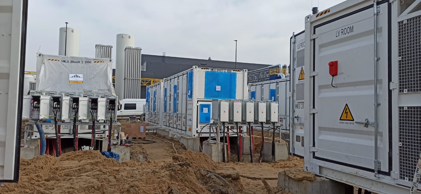 Instalacja kontenerowa z modułami elektrycznymi i zbiornikami kriogenicznymi w tle, teren budowy z piaskiem i elementami instalacji.