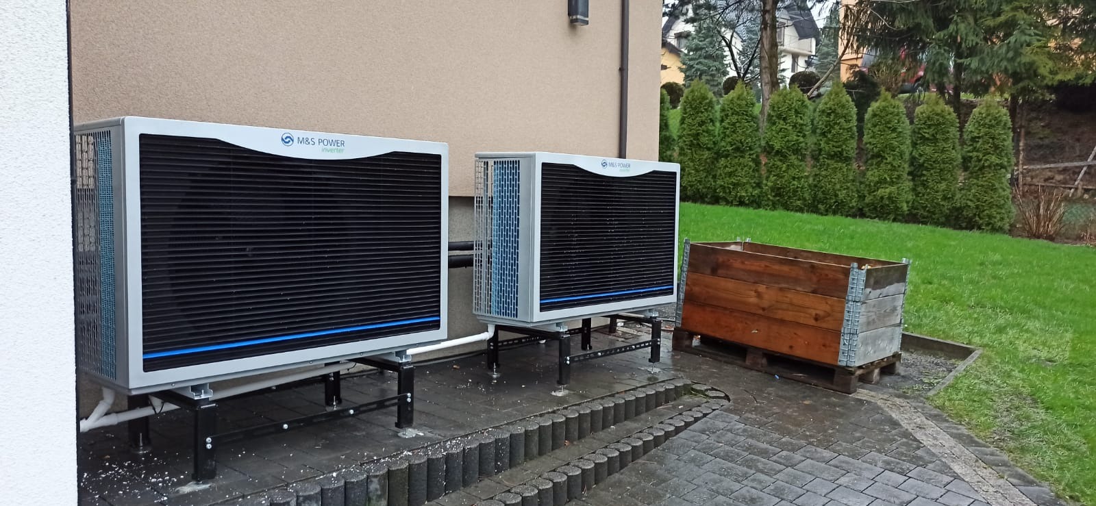 Dwa moduły pompy ciepła powietrze-woda marki M&S Power Inverter zainstalowane na zewnątrz budynku, na szarym brukowanym patio, z drewnianą skrzynią obok, na tle trawnika i iglaków.