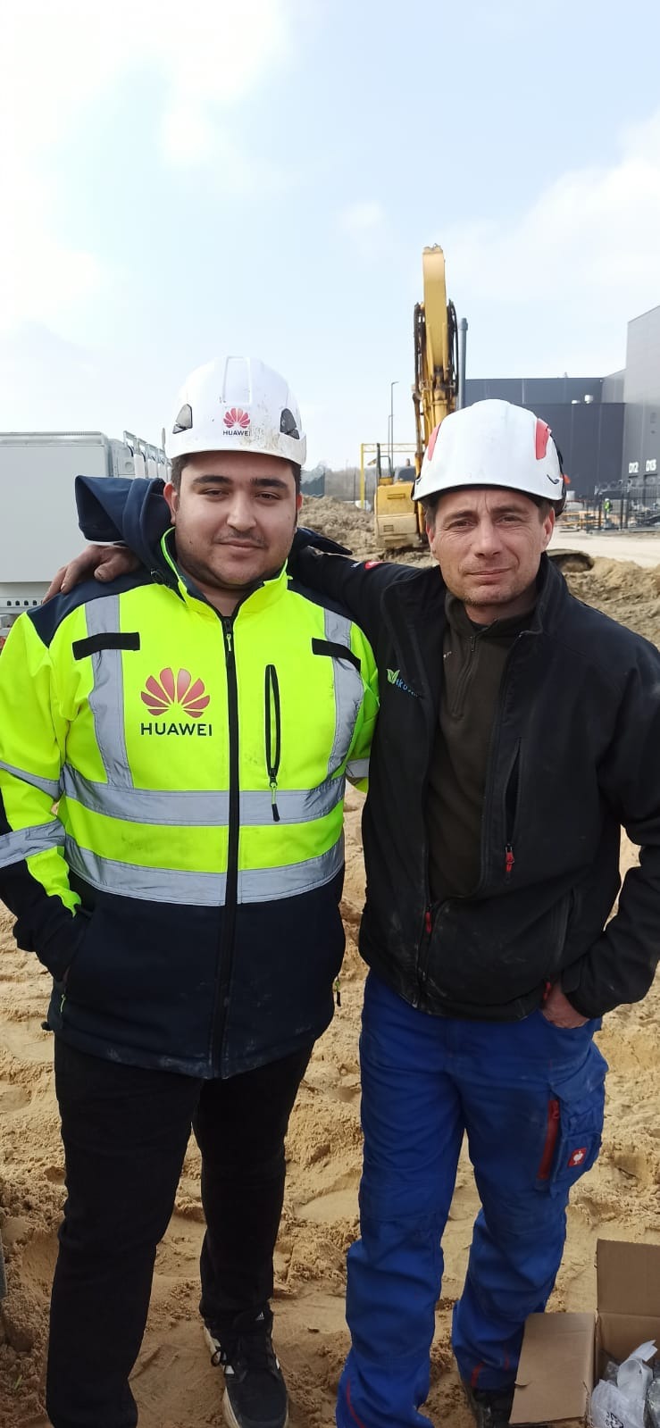 Dwóch inżynierów w kaskach i odzieży roboczej, z logo Huawei na kasku i kurtce, pozuje na placu budowy z koparką w tle, prawdopodobnie po zakończonej realizacji inwestycji.