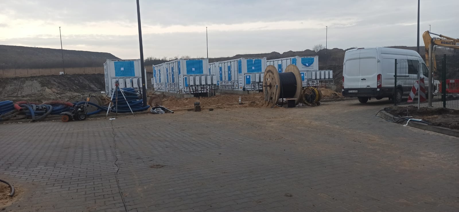 Plac budowy z kontenerami z niebieskimi panelami, bębnami z kablami, rurami, agregatem, białym vanem i koparką w tle.