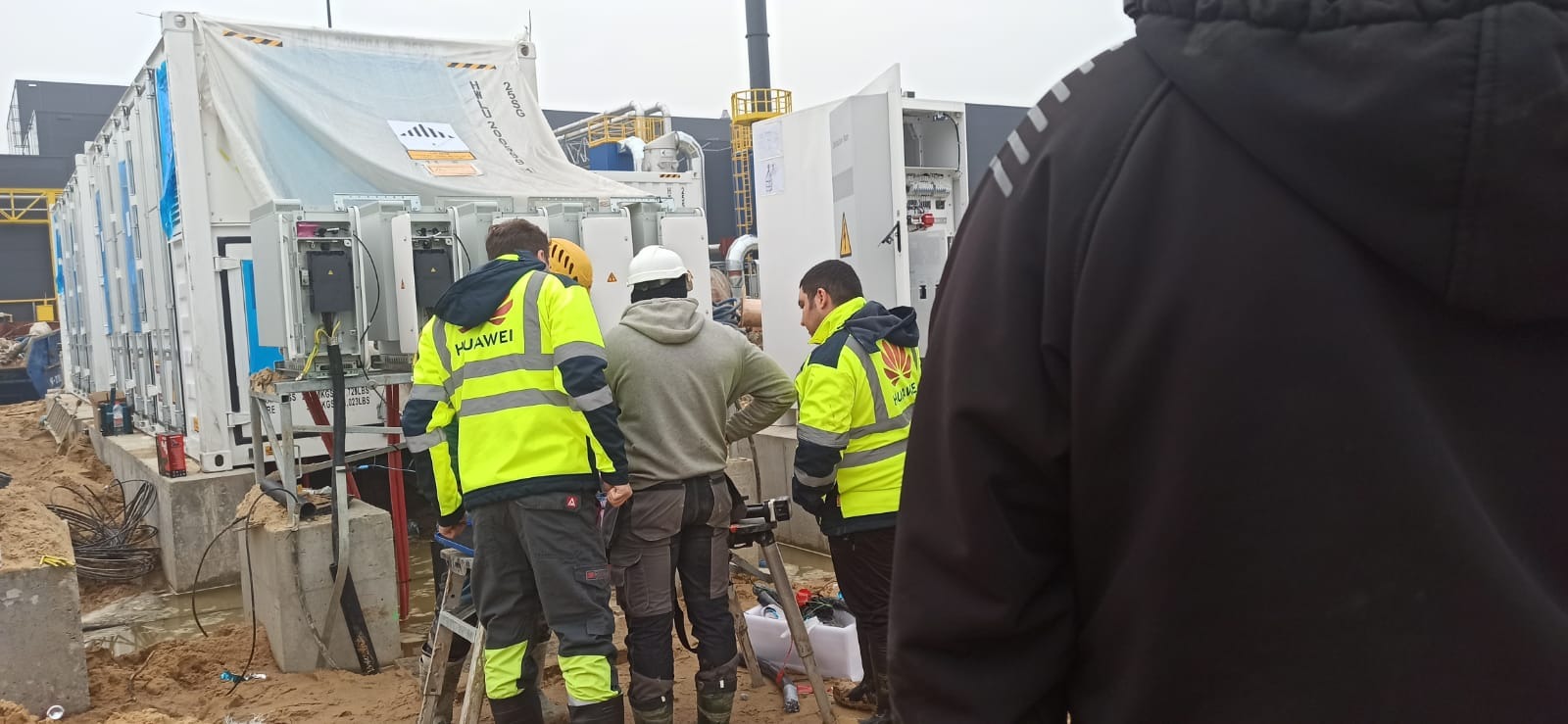 Trzech monterów w odblaskowych kamizelkach Huawei instaluje moduł magazynu energii na betonowych fundamentach w terenie budowy, widoczny częściowo czarny budynek w tle.