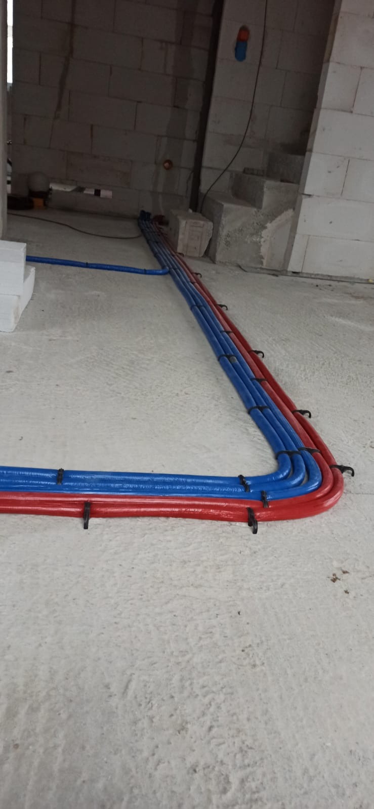 Równolegle ułożone rury instalacyjne w kolorze niebieskim i czerwonym, przymocowane do podłoża czarnymi uchwytami, na tle surowych ścian z betonu komórkowego w trakcie budowy.