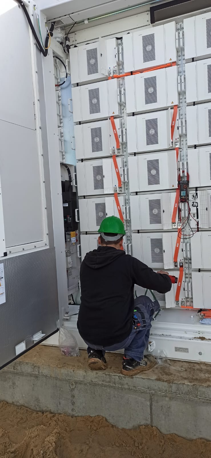Pracownik w zielonym kasku instaluje moduły w szafie magazynu energii, widoczne pomarańczowe pasy mocujące i miernik cęgowy na jednym z modułów.