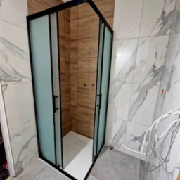 Nowoczesna kabina prysznicowa z czarnymi profilami i matowym szkłem, otoczona płytkami imitującymi drewno i marmur, z widocznymi przewodami elektrycznymi na ścianie.