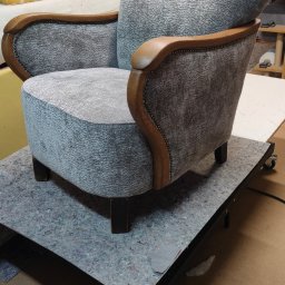 Renowacje Tapicerskie ZDZISŁAW SYCHOWSKI - Odrestaurowany fotel z drewnianymi podłokietnikami i szarą tapicerką o wyraźnej fakturze, umieszczony na wózku warsztatowym w pomieszczeniu z elementami wyposażenia warsztatu.