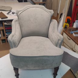 Renowacje Tapicerskie ZDZISŁAW SYCHOWSKI - Odrestaurowany fotel tapicerowany w odcieniach szarości na kółkach, widoczny w warsztacie tapicerskim z maszynami do szycia i wykrojami w tle.