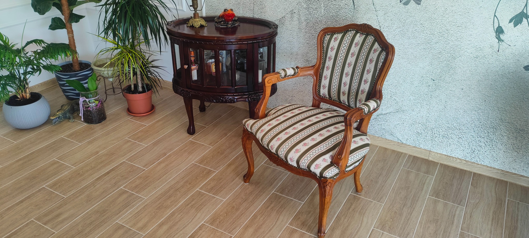 Odrestaurowany fotel w stylu vintage z drewnianą ramą i tapicerką w paski z motywem kwiatowym, stojący na kafelkach w pomieszczeniu z roślinami doniczkowymi i barkiem na alkohol.