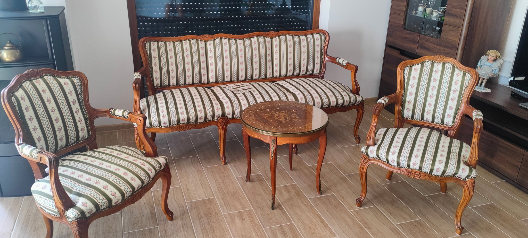 Komplet wypoczynkowy w stylu retro: sofa i dwa fotele z drewnianymi ramami i tapicerką w paski z motywem kwiatowym, uzupełniony okrągłym stolikiem z blatem inkrustowanym kwiatami.