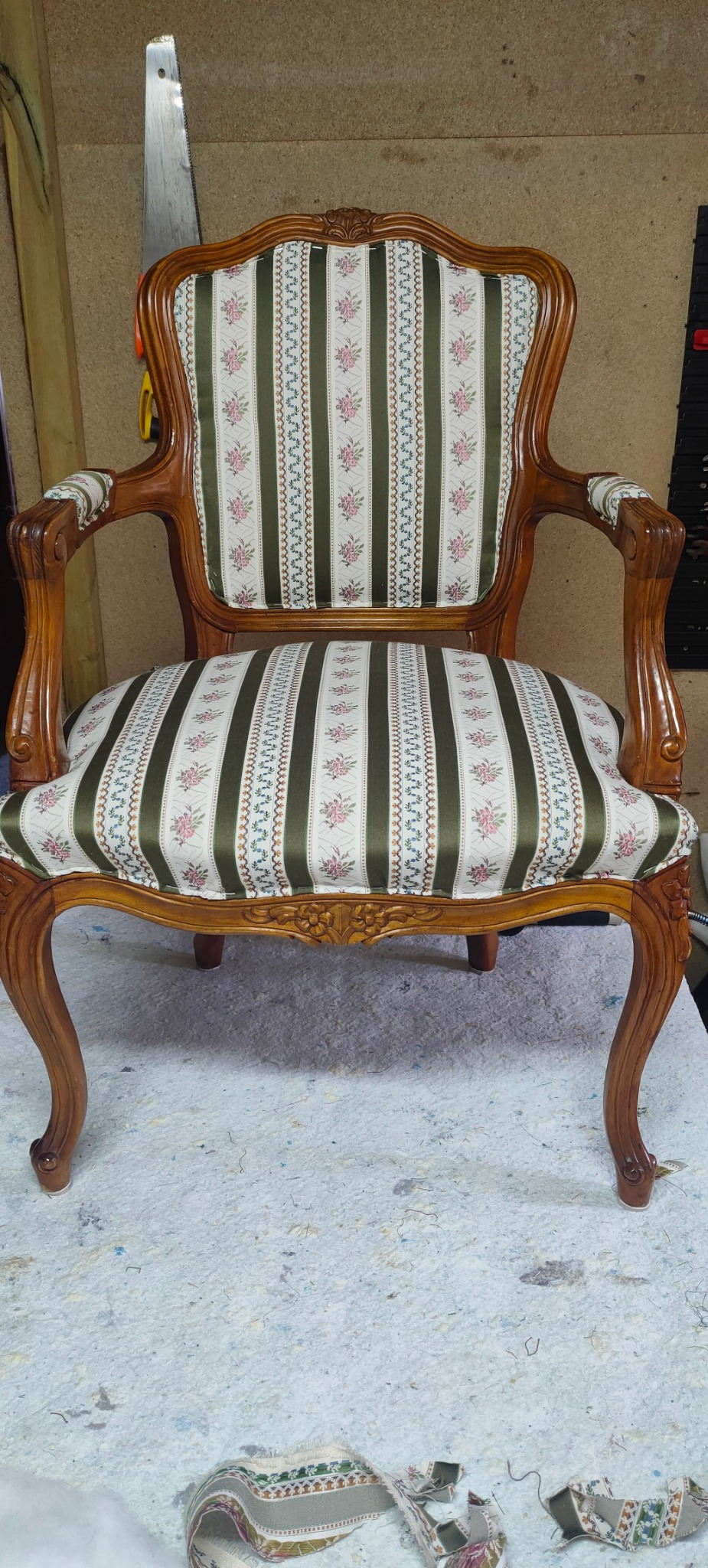 Odrestaurowany fotel w stylu Ludwika Filipa z nową tapicerką w zielono-białe pasy z motywem kwiatowym, stojący na powierzchni roboczej z resztkami materiałów.