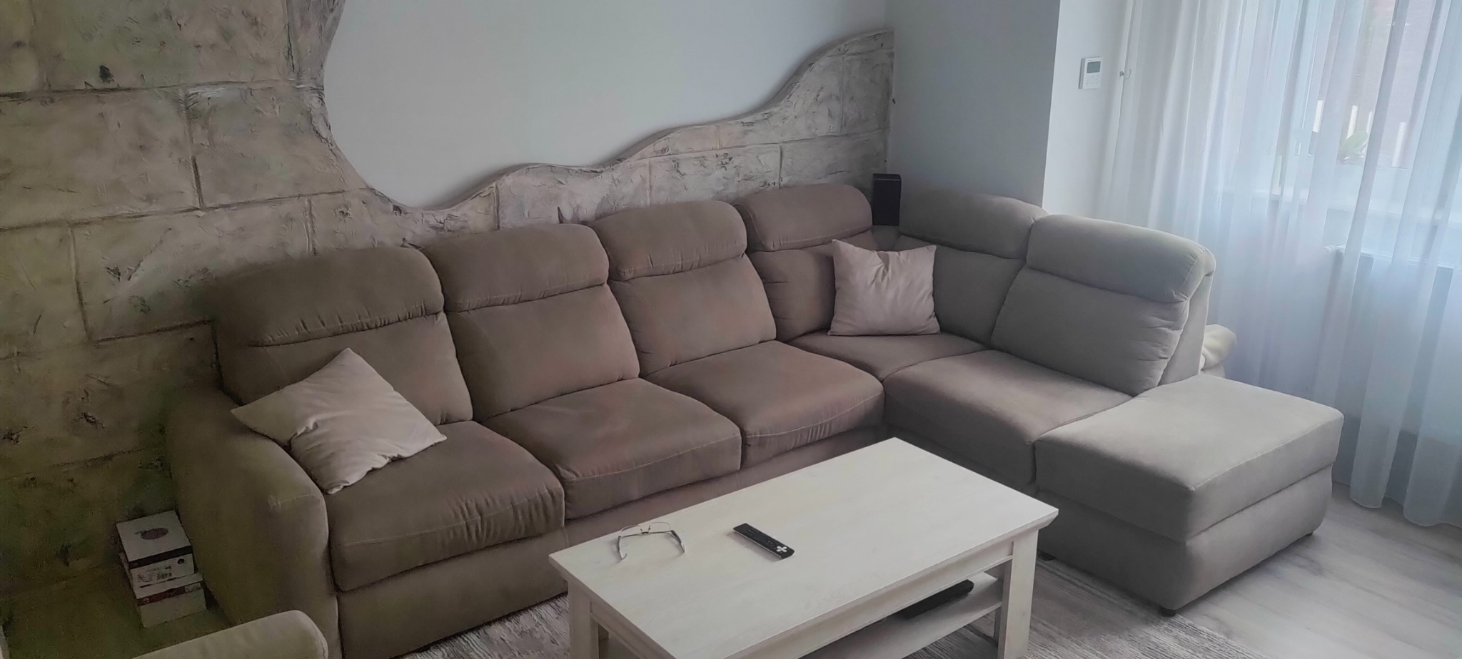 Beżowy narożnik z poduszkami w salonie z dekoracyjną ścianą imitującą kamień, obok okno z białymi firankami, na stoliku okulary i pilot.