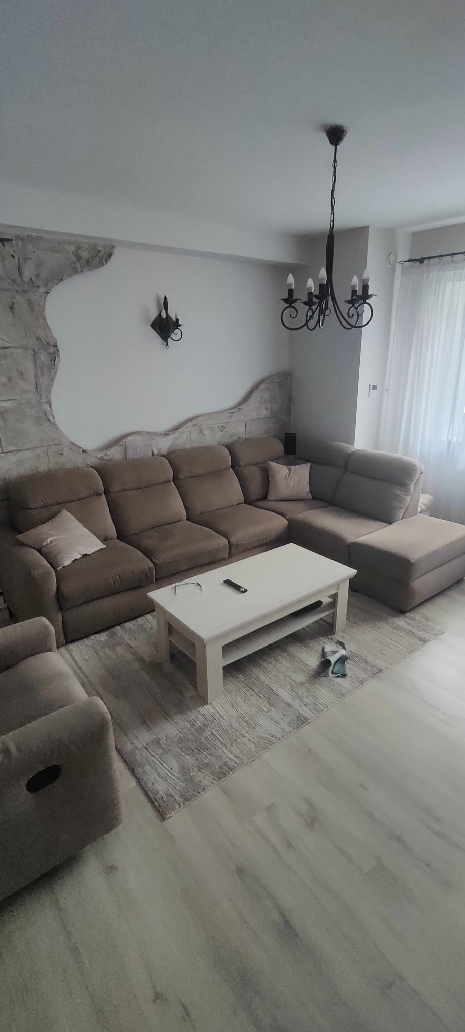 Przestronny salon z narożną sofą w odcieniach beżu, białym stolikiem kawowym z okularami i pilotem, oraz dekoracyjną ścianą imitującą kamień. W tle widoczny żyrandol i zasłony.