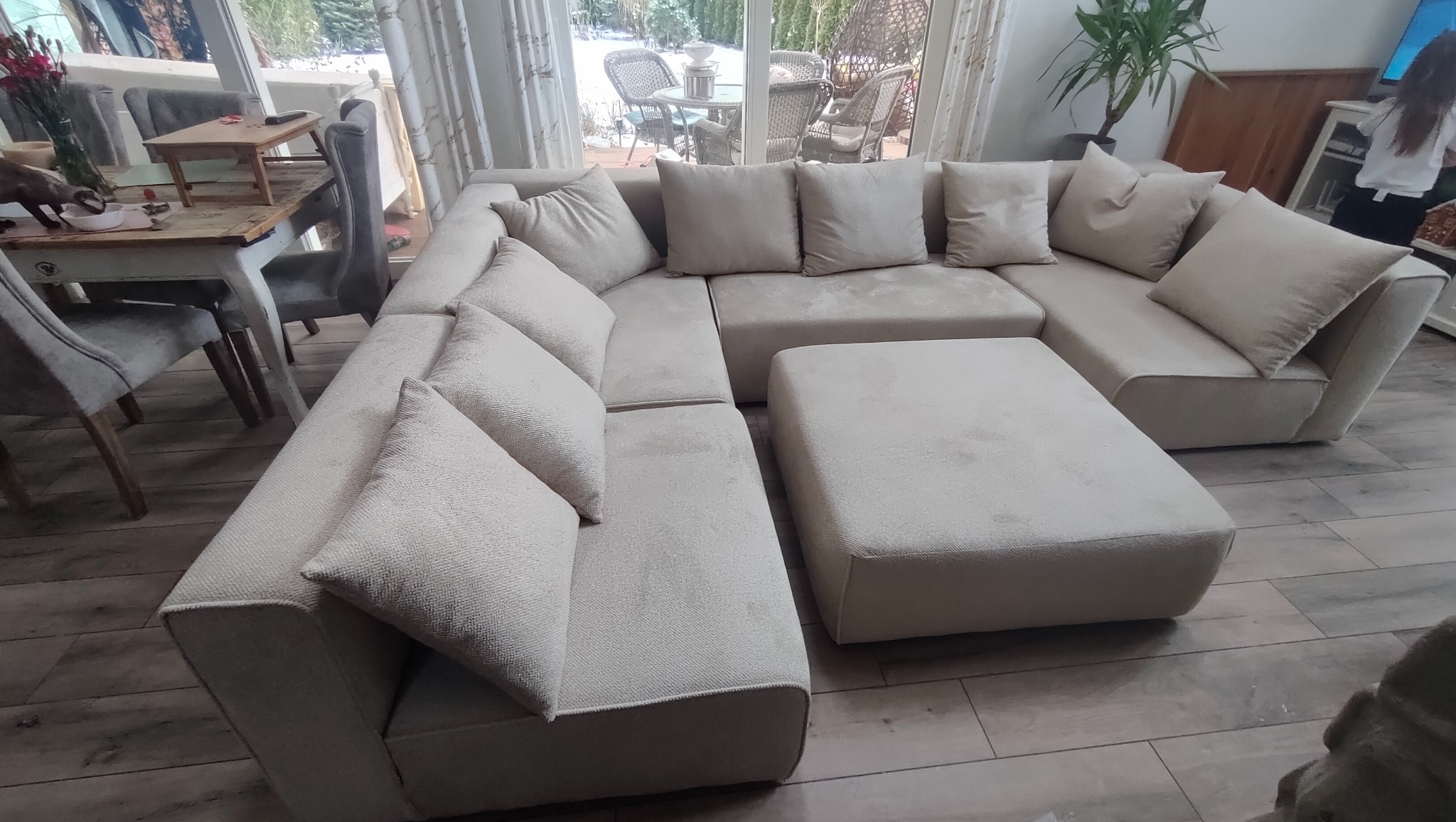 Jasny, modułowy narożnik z pufą i poduszkami w salonie z drewnianą podłogą, widok na ogród przez okno.