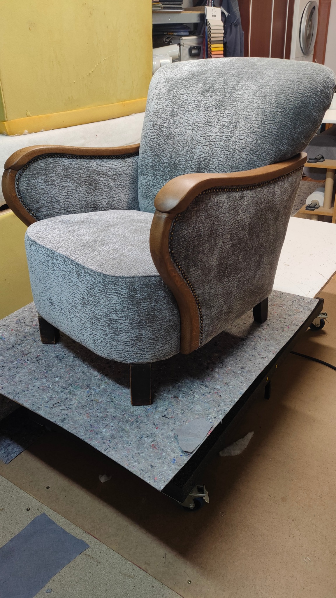 Odrestaurowany fotel z drewnianymi podłokietnikami i szarą tapicerką o wyraźnej fakturze, umieszczony na wózku warsztatowym w pomieszczeniu z elementami wyposażenia warsztatu.