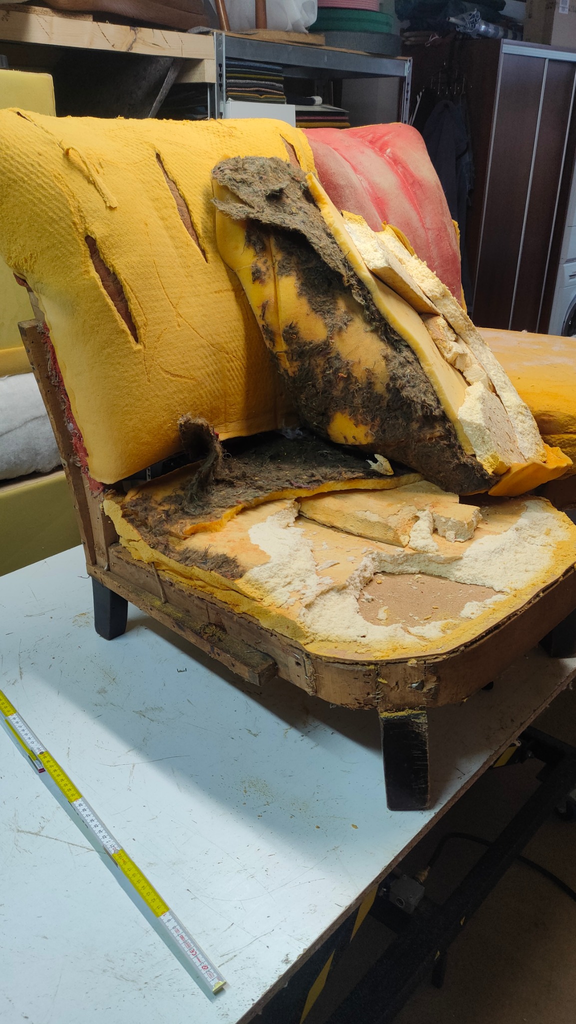Zniszczony fotel z odsłoniętą konstrukcją, żółtą pianką i resztkami starej tapicerki na stole warsztatowym, widoczna miarka.