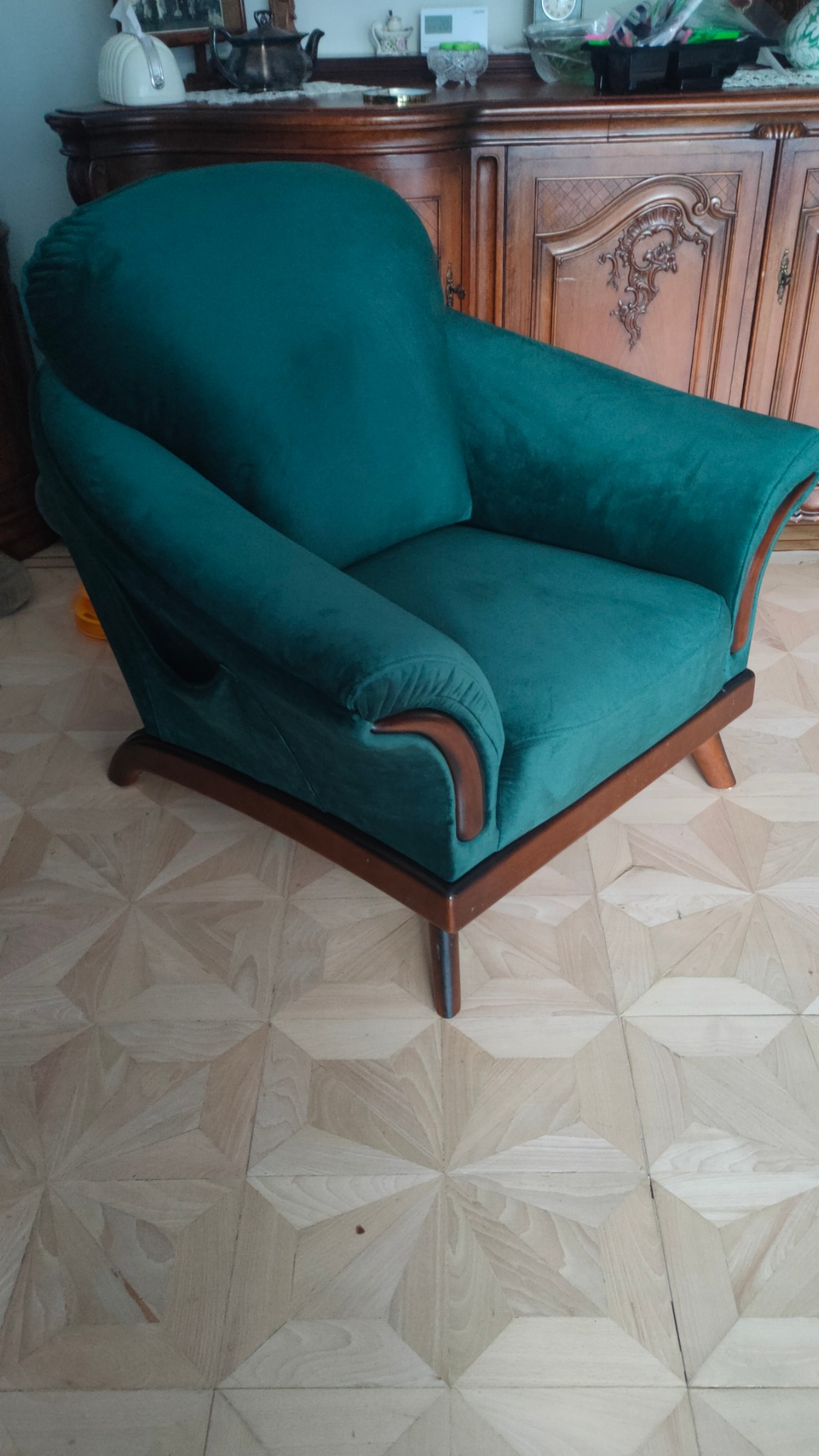 Szmaragdowy fotel klubowy z drewnianymi podłokietnikami stoi na parkiecie w geometryczny wzór, w tle widoczny kredens.