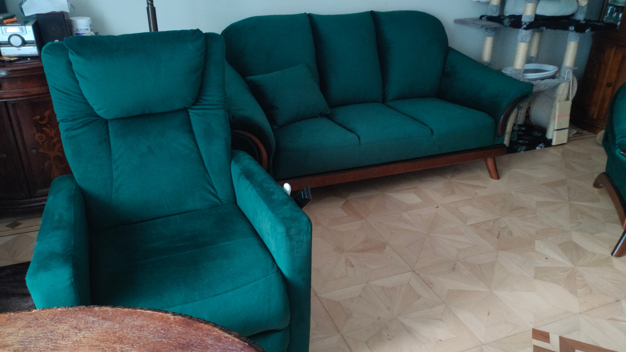 Szmaragdowy fotel z wysokim oparciem i sofa z poduszką, po renowacji, na parkiecie w jodełkę.