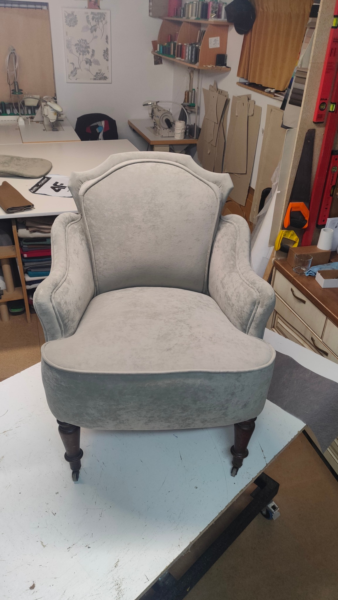 Odrestaurowany fotel tapicerowany w odcieniach szarości na kółkach, widoczny w warsztacie tapicerskim z maszynami do szycia i wykrojami w tle.