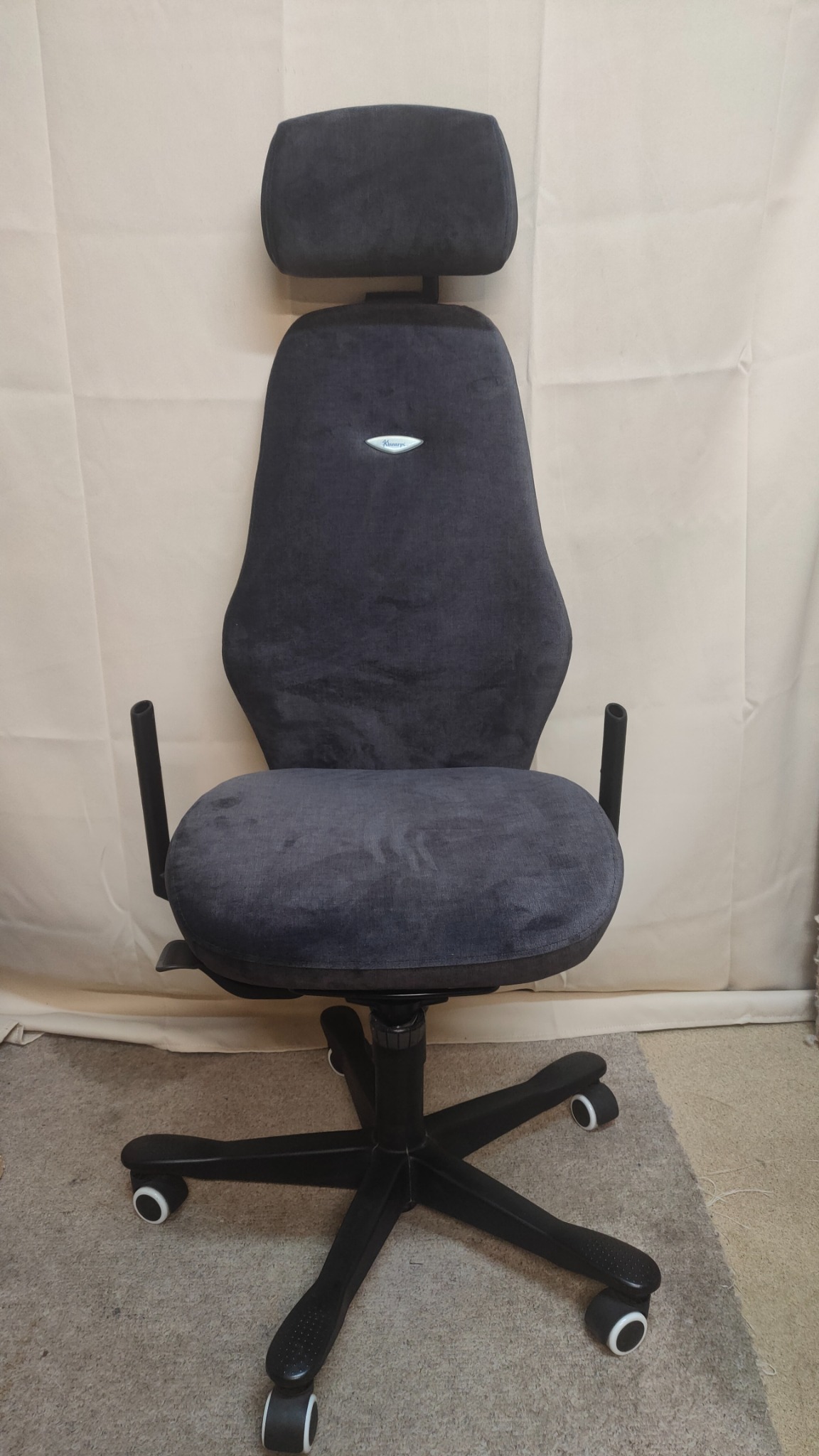 Ergonomiczne, obite szarą tkaniną krzesło biurowe z zagłówkiem, na czarnej podstawie z kółkami, ustawione na dywanie.