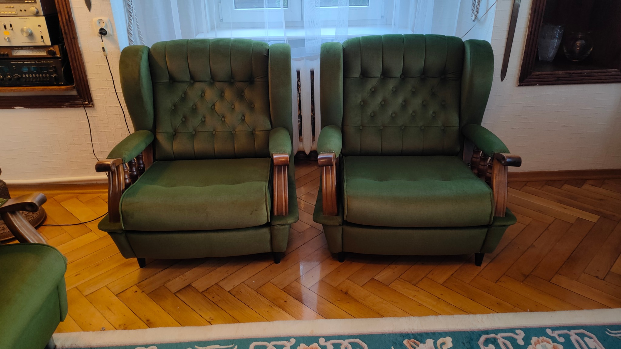 Dwa zielone, tapicerowane fotele uszak z drewnianymi podłokietnikami, ustawione obok siebie na parkiecie, z widocznymi guzikami na oparciach i elementami starego sprzętu audio w tle.