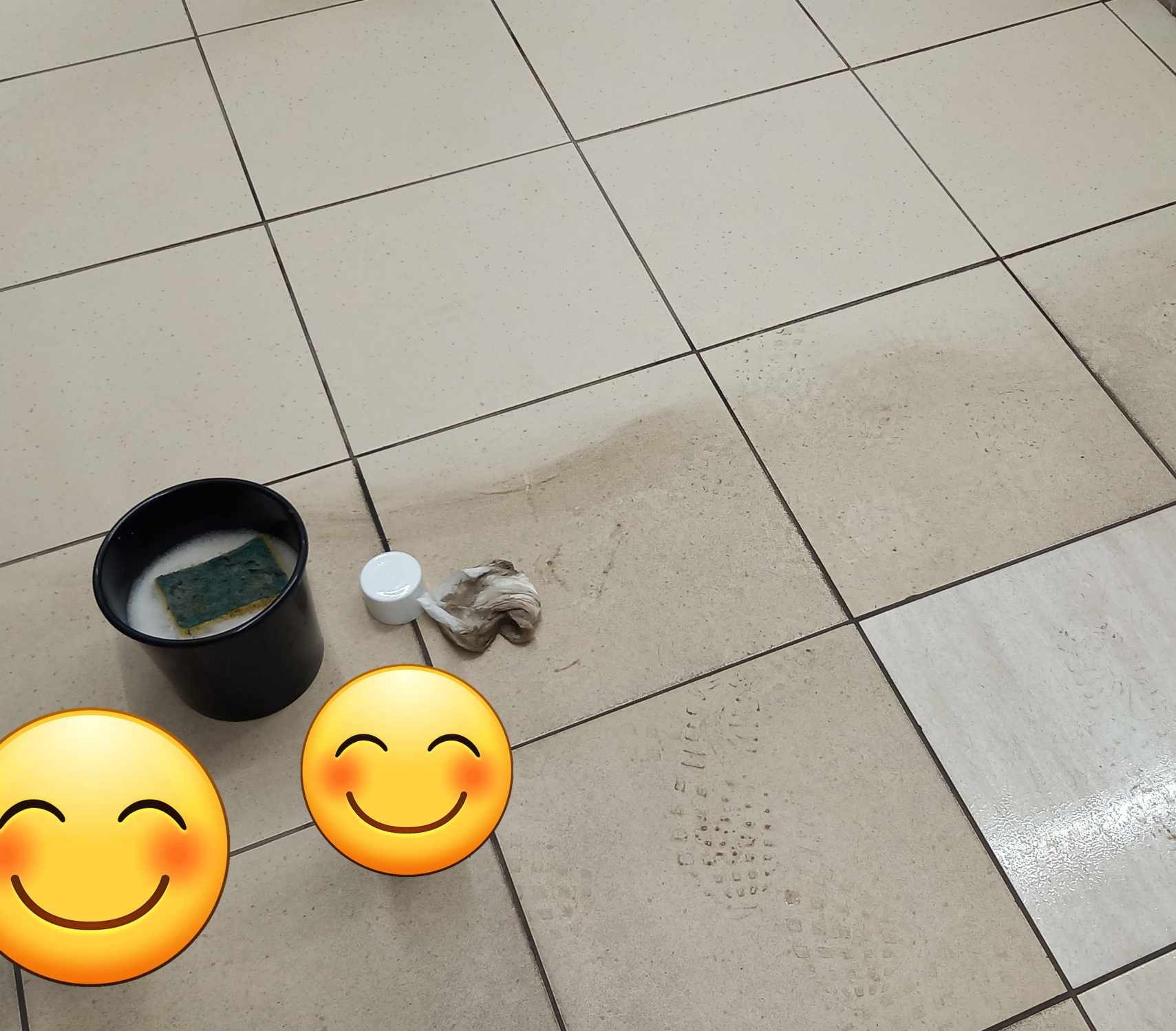 Czyszczona podłoga z płytek ceramicznych, widoczny czarny pojemnik z wodą i gąbką, brudna szmatka, plama i ślady butów na płytkach.