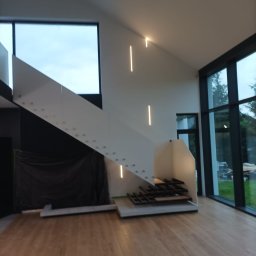 GENERAL HELP Sp.z.o.o - Wnętrze domu w trakcie budowy, widoczne nowoczesne schody z białymi panelami, duże okna z widokiem na zieleń, drewniana podłoga i minimalistyczne oświetlenie liniowe.