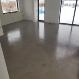 Wnętrze w stanie deweloperskim z wylewką samopoziomującą na podłodze i dużymi oknami z widokiem na zimowy krajobraz.