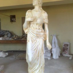 GENERAL HELP Sp.z.o.o - Biała, marmurowa statua kobiety w drapowanej szacie, ustawiona w warsztacie rzeźbiarskim z widocznymi narzędziami i elementami w trakcie obróbki.