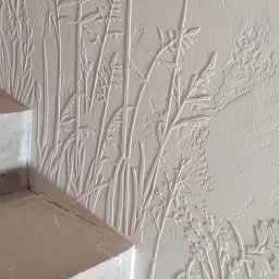 GENERAL HELP Sp.z.o.o - Wypukła dekoracja ścienna w odcieniach bieli, przedstawiająca motyw roślinny w pobliżu schodów, z widoczną fakturą tynku.