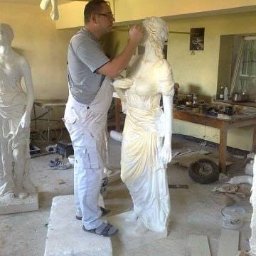 GENERAL HELP Sp.z.o.o - Artysta w szarym t-shircie i białych spodniach roboczych retuszuje gipsową statuę kobiety w pracowni, widoczne inne rzeźby i narzędzia.